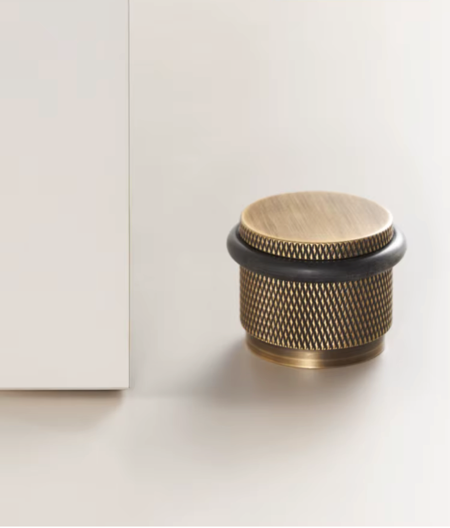 Solid brass knurled floor door stop in antique brass with a black rubber ring beside a white door